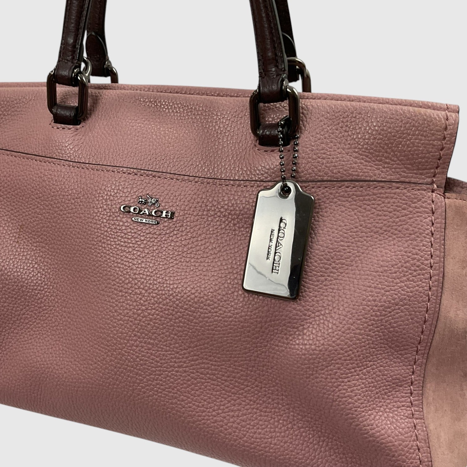 A close-up of a Coach handbag in dusty pink pebbled leather with dark brown leather handles and a silver-tone metal logo plate and hangtag.