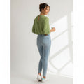 Woman wearing a green top and light blue jeans holding a mug in a bright room.