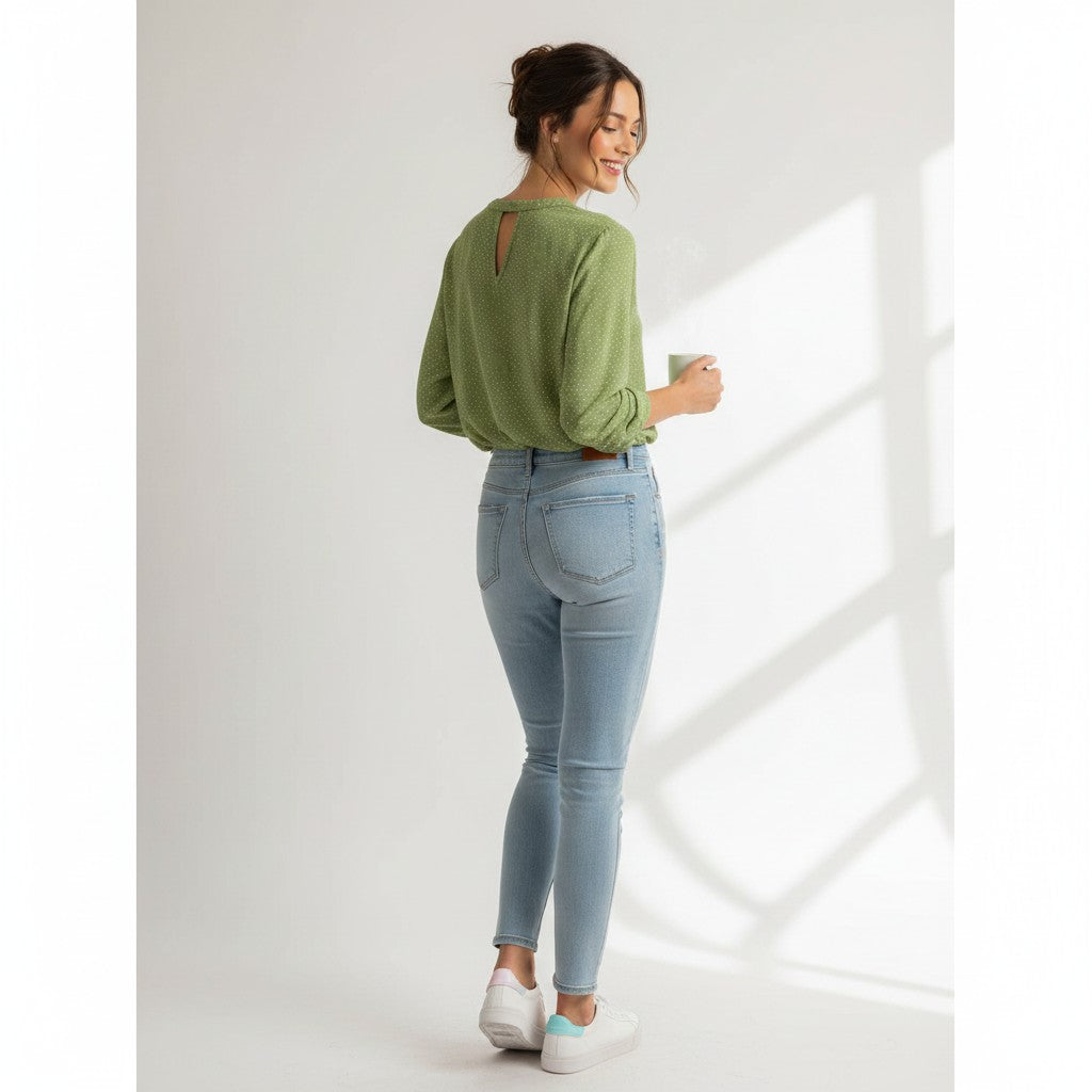 Woman wearing a green top and light blue jeans holding a mug in a bright room.