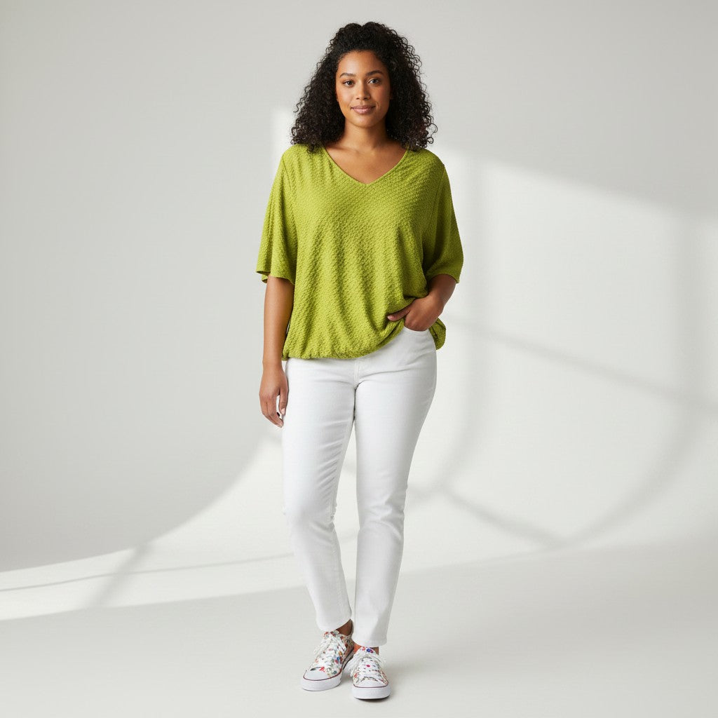 A woman modeling a textured lime green short-sleeve V-neck top with white pants and white sneakers, standing against a light gray studio background with soft shadows.
