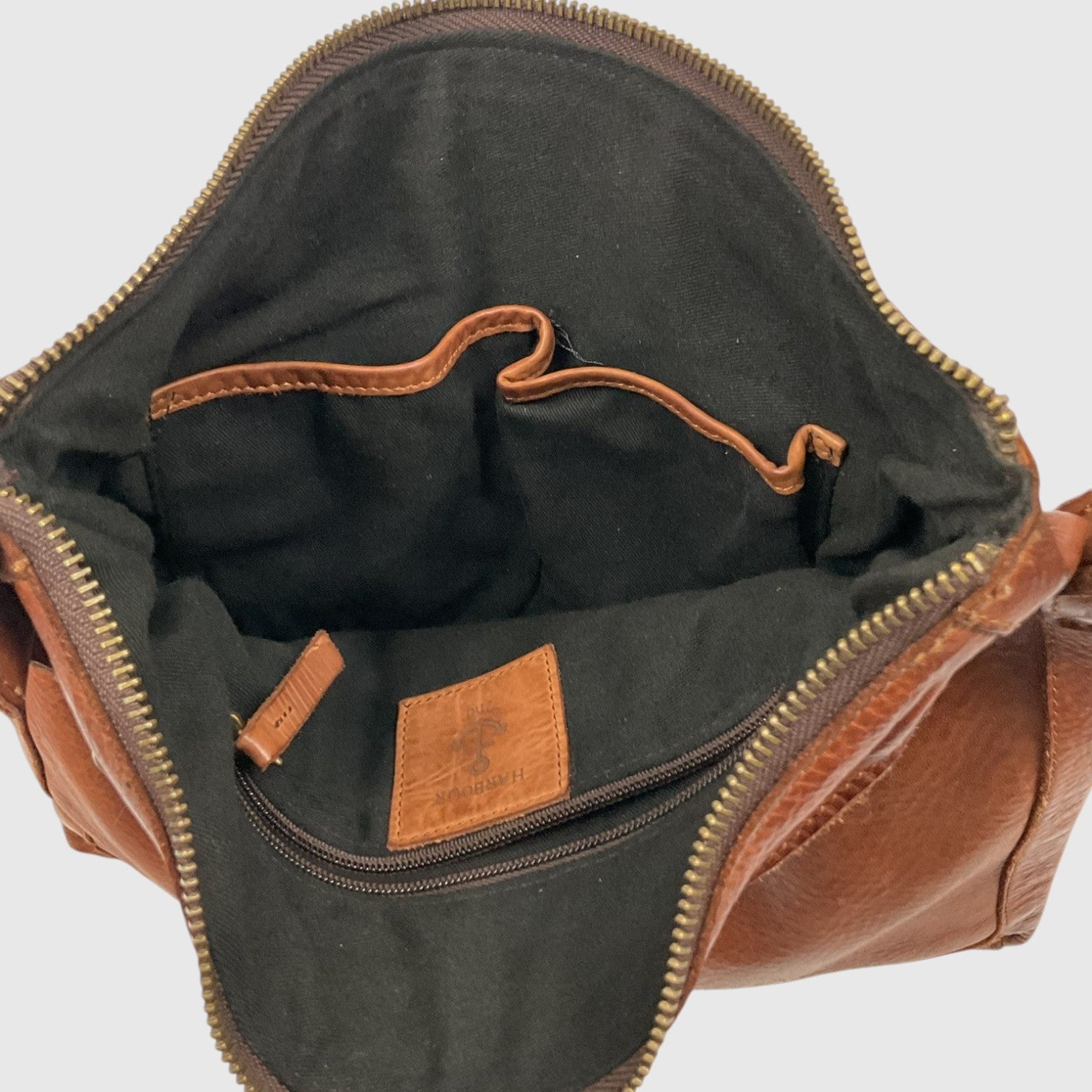 Close-up of the interior of a brown leather bag, showing a black fabric lining, gold-toned zipper, interior pocket, and a small tan leather brand patch.