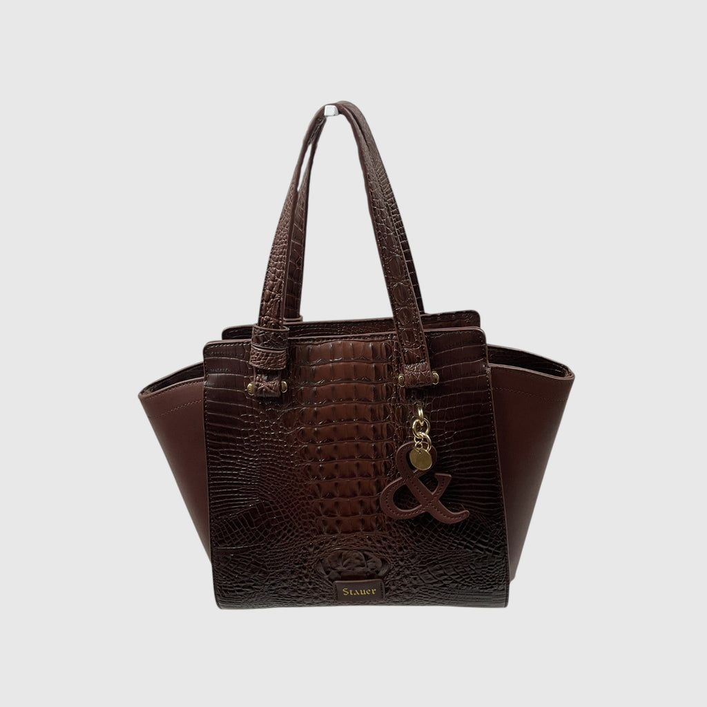A dark brown tote bag with a crocodile-embossed front panel and smooth brown leather side panels and handles, featuring a gold-tone charm with an ampersand symbol and a small 'STUART' logo on the front, set against a light gray background.