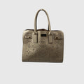 A structured taupe leather handbag with laser-cut floral patterns, dual top handles, and gold-tone hardware, shown against a light gray background.