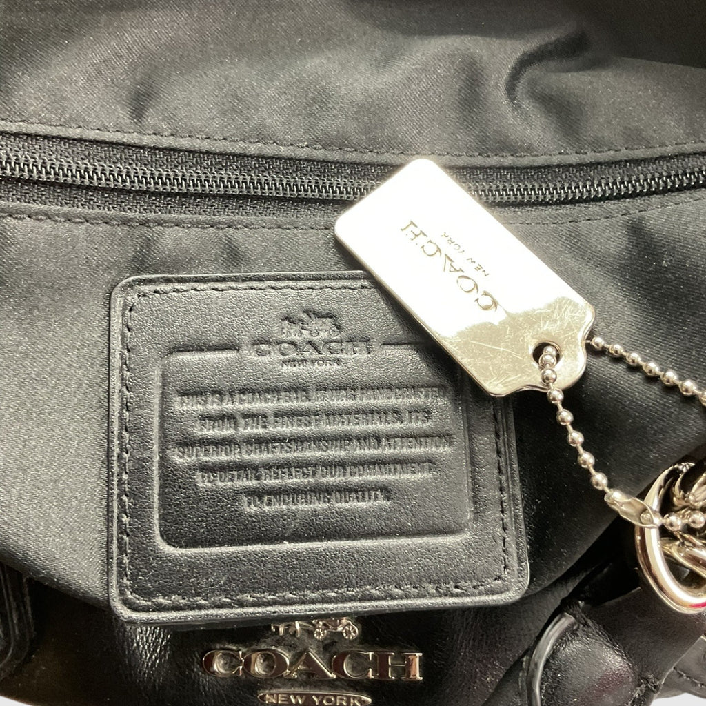 Close-up of a Coach bag showing a black leather patch with embossed branding, a silver-tone metal hangtag, and part of a gold-tone logo charm on a black fabric background.
