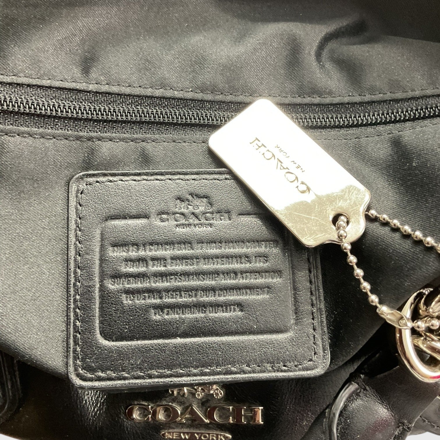 Close-up of a Coach bag showing a black leather patch with embossed branding, a silver-tone metal hangtag, and part of a gold-tone logo charm on a black fabric background.