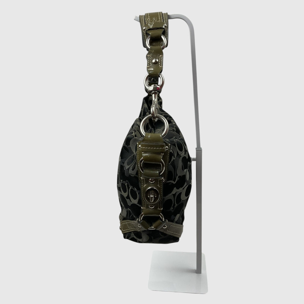 A small black and gray patterned handbag with olive green leather trim and silver-tone hardware, displayed on a white metal stand against a light gray background.
