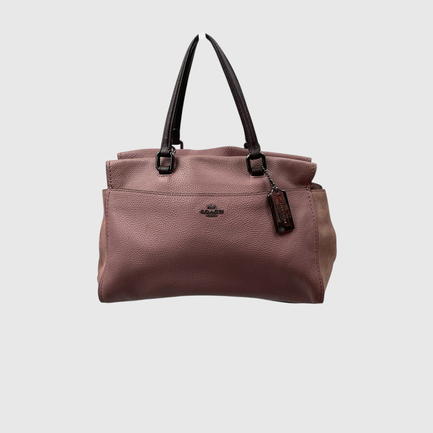 A Coach pebbled leather handbag in dusty rose pink, featuring dark brown leather top handles and a small metal logo plaque on the front, set against a solid light gray background.