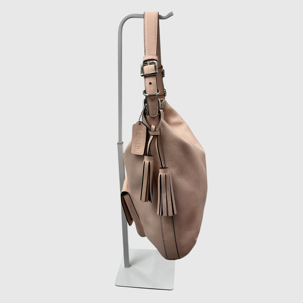 A taupe leather slouchy shoulder bag with tassel details and a buckle strap, hanging from a minimalist gray display stand against a light gray background.