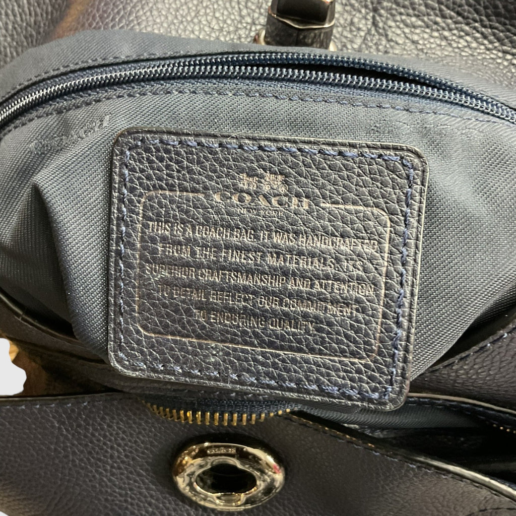 Close-up of a Coach handbag interior showing a dark gray pebbled leather patch with embossed Coach branding and text, along with zipper and metal hardware.