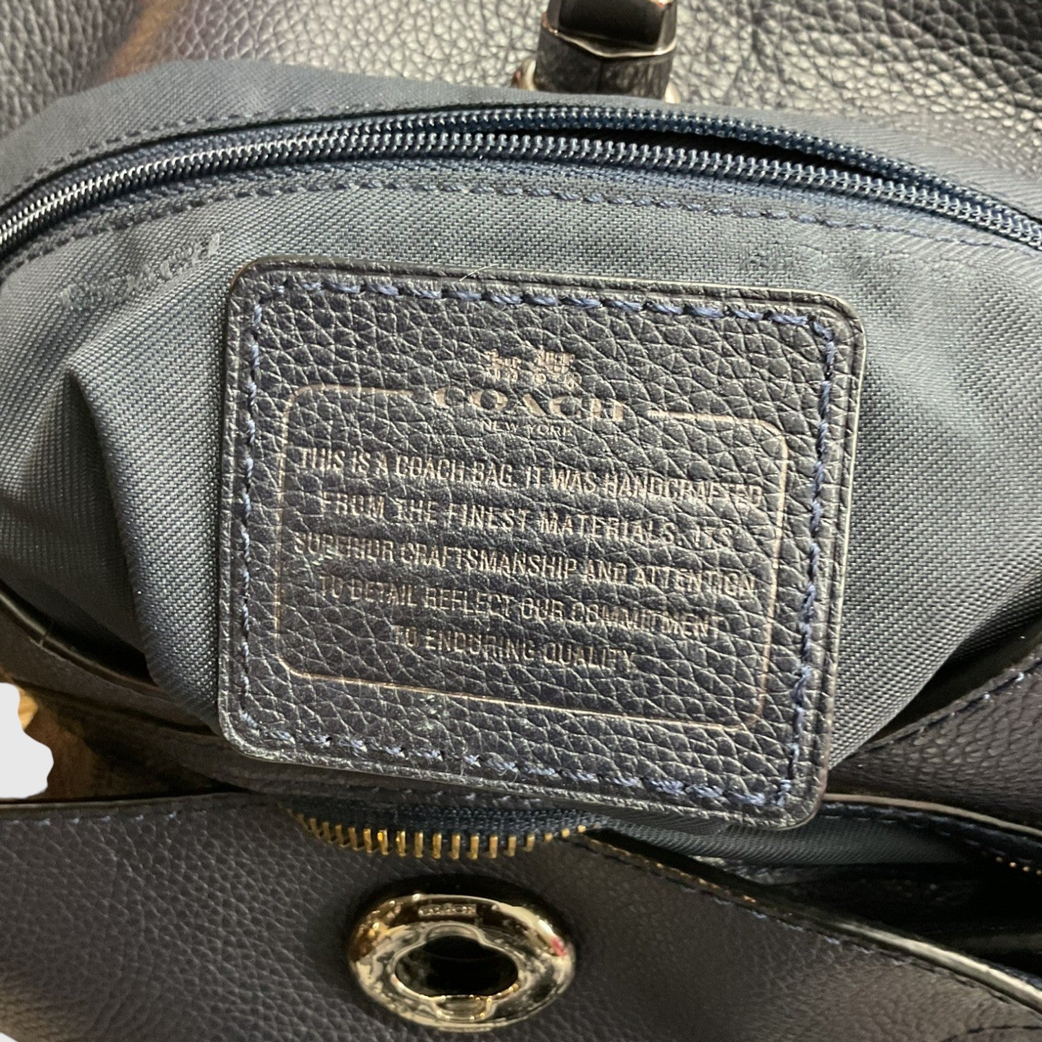 Close-up of a Coach handbag interior showing a dark gray pebbled leather patch with embossed Coach branding and text, along with zipper and metal hardware.