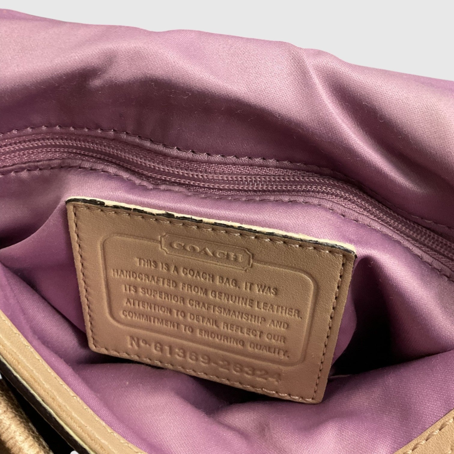 Close-up of the interior of a Coach bag showing a tan leather brand patch sewn onto a lavender satin-lined compartment with a zipper.