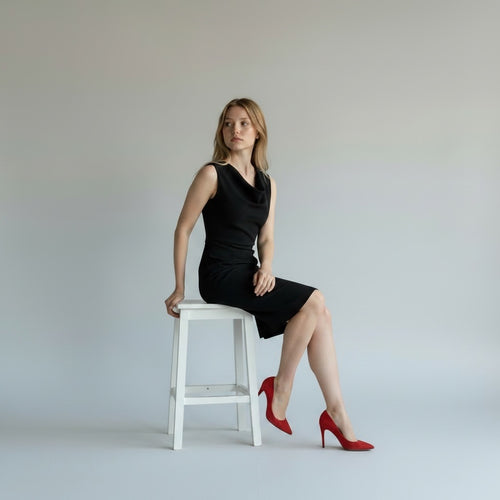 Woman in a black dress and red shoes sitting on a white stool against a plain background