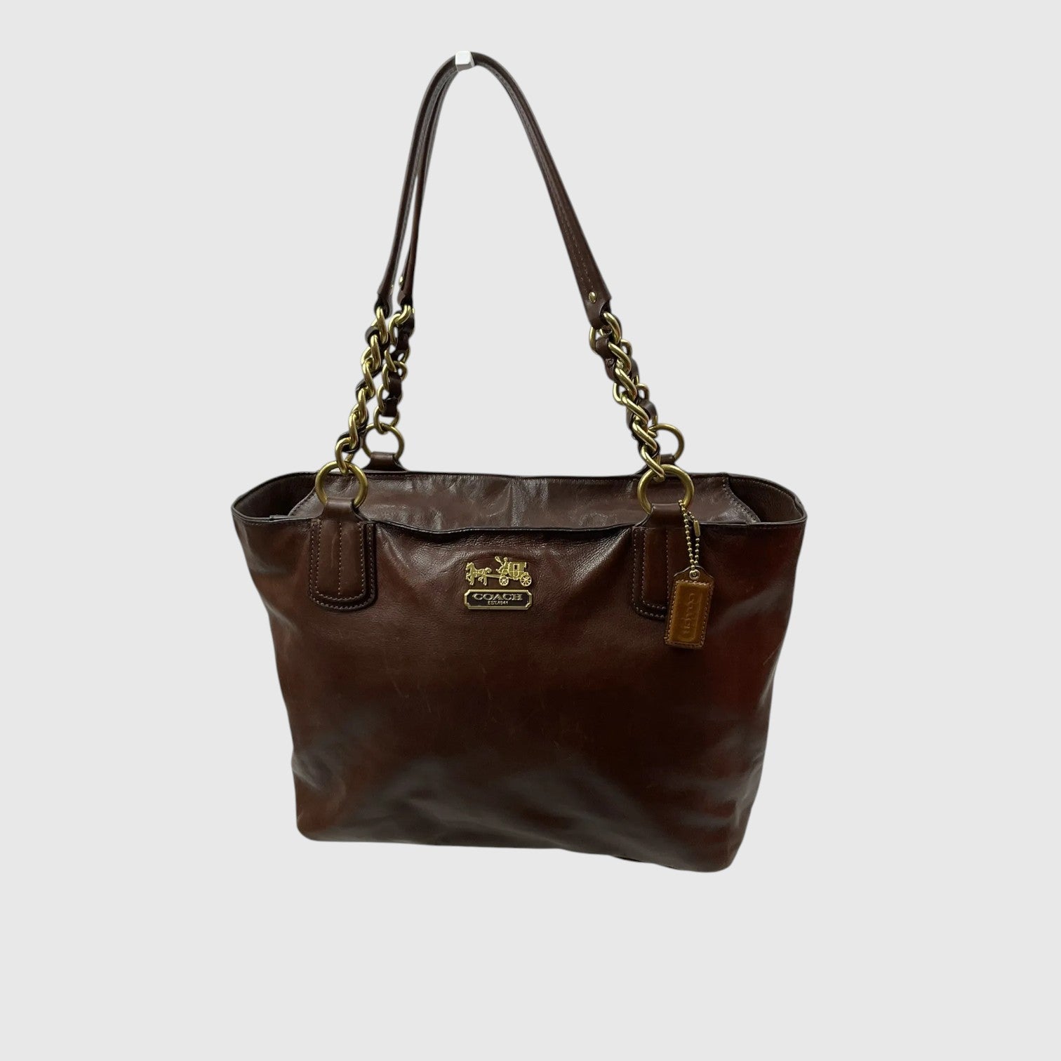 A dark brown leather tote bag with gold-tone hardware, featuring a gold-tone Coach logo plaque on the front and a leather hangtag, set against a plain light gray background.